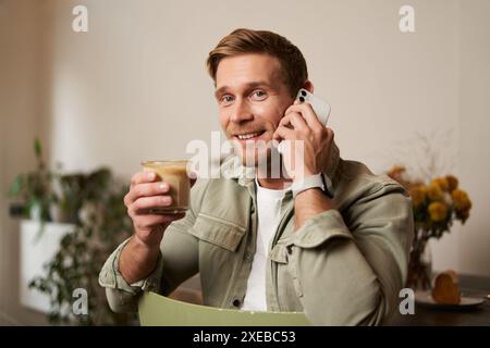 Bild eines lächelnden, gut aussehenden jungen blonden Mannes mit Telefon, Kaffee im Café trinken, mit jemandem telefonieren, Antwort Stockfoto