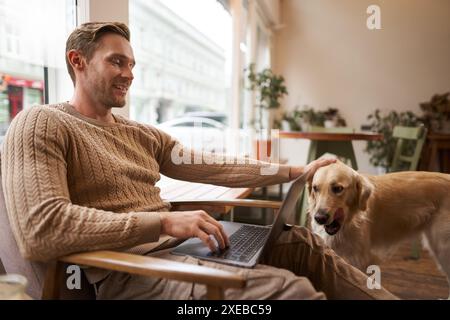 Hübscher junger Mann, der mit einem Hund in einem Café arbeitet, auf einem Stuhl sitzt und einen Laptop benutzt, seinen goldenen Retriever tierfreundlich streichelt Stockfoto