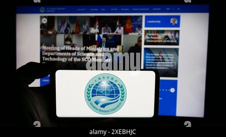 Person, die ein Mobiltelefon mit dem Logo der Eurasischen Shanghai Cooperation Organisation (SCO) vor der Webseite hält. Schwerpunkt auf dem Display des Telefons. Stockfoto