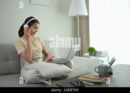 Glückliche junge asiatische Frau in kabellosen Kopfhörern, die zu Hause auf einem Laptop Filme ansieht Stockfoto