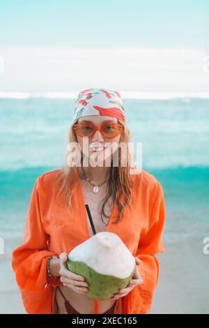 Porträt einer bezaubernden rothaarigen Frau in orangefarbenem Hemd hält Kokosnuss in ihren Händen Stockfoto