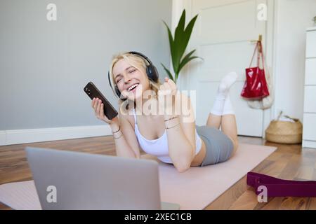 Junge Frau, die zu Hause Fitness-Workout macht, auf einer Gummi-Yoga-Matte liegt, Smartphone hält, Kopfhörer trägt, Laptop benutzt, um zu waten Stockfoto