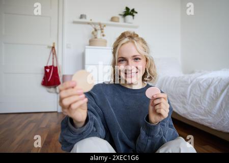 Bild einer lächelnden blonden Frau, Vloggerin, die Make-up-Produkte, Schönheitsartikel zeigt, Followern empfiehlt, auf dem Boden im Bett sitzen Stockfoto