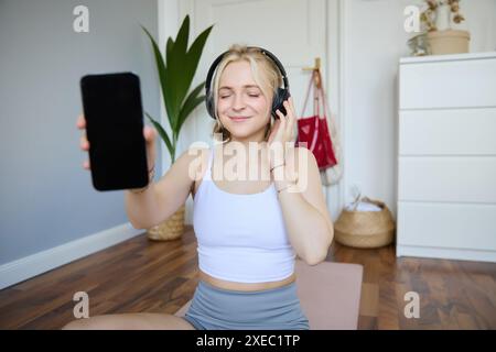 Porträt einer lächelnden, schönen jungen Frau, die ihren Smartphone-Bildschirm zeigt, Kopfhörer trägt und mit der Klangqualität zufrieden aussieht Stockfoto