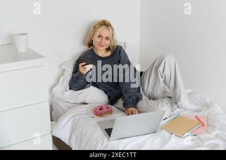 Porträt einer jungen Frau, Studentin, die in ihrem Bett studiert, sich entspannt, während sie Hausaufgaben vorbereitet, Donut isst, Laptop im Schlafzimmer benutzt Stockfoto