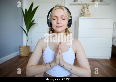 Porträt einer Frau, die zu Hause meditiert, in Kopfhörern sitzt, Yoga-Podcasts hört, Hände im Namensschild hält Stockfoto