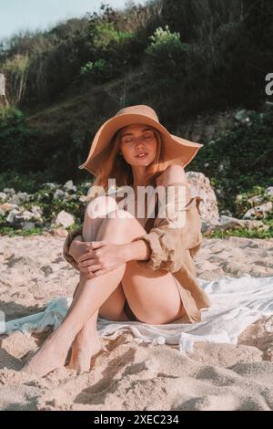 Junge attraktive rothaarige Frau mit Strohhut und braunem Bikini sitzt am Strand Stockfoto