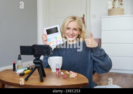 Porträt einer jungen lächelnden Frau, eines Mädchens, das den Lidschatten überprüft, Daumen hoch zeigt, Video-Blog aufnimmt, Vloggen in ihrem Zimmer, Stockfoto