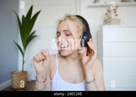 Porträt der glücklichen Frau, hört Musik in kabellosen Kopfhörern, hält unsichtbares Mikrofon, singt ins Mikrofon, lächelt, genießt Stockfoto