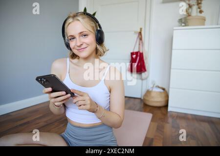 Nahaufnahme des Porträts einer sportlichen jungen Frau, die ihr Mobiltelefon hält und schnurlose Kopfhörer trägt, zu Hause trainieren, mit Sport ap Stockfoto