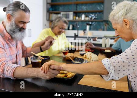 Zu Hause, eine Gruppe von älteren Freunden, die Hände halten und vor dem Essen in der Küche beten Stockfoto