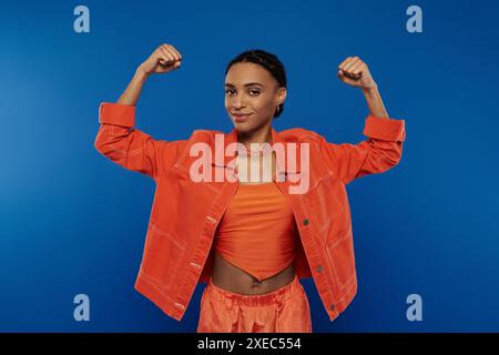 Eine hübsche junge Afroamerikanerin zeichnet sich in einer leuchtend orangefarbenen Jacke und Shorts vor blauem Hintergrund aus. Stockfoto