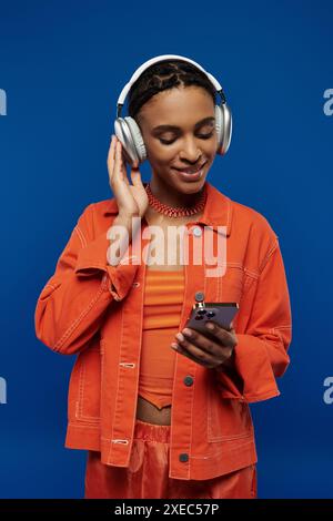 Junge afroamerikanische Frau in orangefarbenem Outfit, mit Kopfhörern, vertieft in Handy-Bildschirm auf blauem Hintergrund. Stockfoto