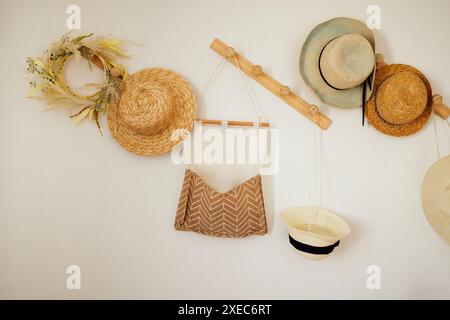 Eine weiße Wand mit Holzbügeln mit verschiedenen Strohhüten. Wandtasche und Kranz aus Baumwollleinen. Stockfoto