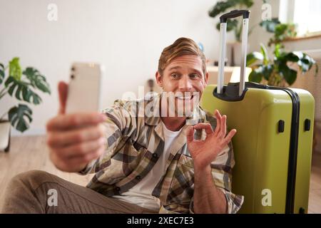 Porträt eines hübschen lächelnden Mannes mit Koffer, nimmt Selfie auf dem Smartphone, zeigt OK-Zeichen, genehmigt Urlaubsziel, postet ph Stockfoto