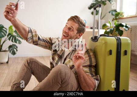 Porträt eines lächelnden, gutaussehenden Mannes mit Koffer, Selfie mit Gepäck, Posts in sozialen Medien über Urlaub Stockfoto