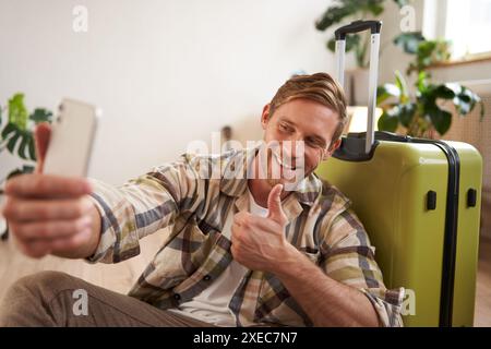 Glücklicher junger Mann zeigt Daumen hoch, macht Selfie auf dem Handy, posiert in der Nähe von Koffern, Blogger geht in den Urlaub und postet über Vaca Stockfoto