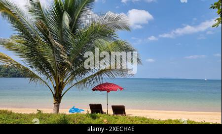 Zwei verwitterte Stühle sitzen unter einem leuchtend roten Sonnenschirm an einem Sandstrand und schaffen einen gemütlichen und einladenden Ort zum Entspannen Stockfoto