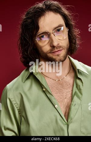 Ein eleganter Mann posiert in Brille und grünem Seidenhemd vor einem leuchtend roten Hintergrund. Stockfoto