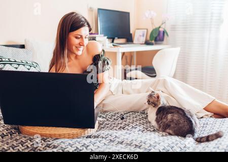 Lächelnde Frau, die von zu Hause aus online mit einem Laptop arbeitet. Die Katze liegt vom Besitzer auf der Couch. Neugieriges Haustier, das auf den Bildschirm schaut. Lifestyle Stockfoto