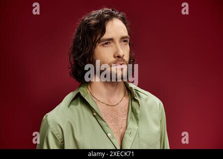Ein gutaussehender Mann mit langen Haaren und Brille posiert in einem grünen Seidenhemd vor einem leuchtend roten Hintergrund. Stockfoto
