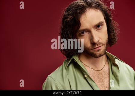 Ein stilvoller Mann mit langen Haaren und einer Brille posiert in einem leuchtend grünen Seidenhemd vor einem leuchtend roten Hintergrund. Stockfoto