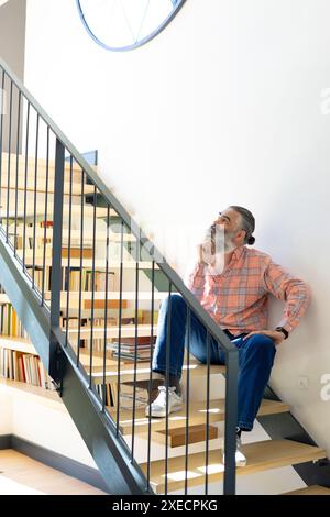 Zu Hause, sitzend auf der Treppe, Seniorenmann in lässigen Kleidern nachdenkend, Bücherregal im Hintergrund Stockfoto