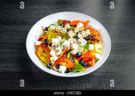 Lebendiger mediterraner Salat mit frischem Gemüse, Fetakäse, schwarzen Oliven und sonnengetrockneten Tomaten, serviert in einer weißen Schüssel auf dunklem Hintergrund Stockfoto