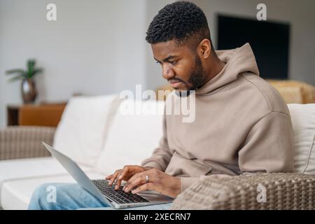 Glücklicher Mann in braunem Hoodie mit Laptop, Lernen, Einkaufen, online arbeiten, ich spreche mit Freunden, während ich zuhause auf einer Couch sitze Stockfoto