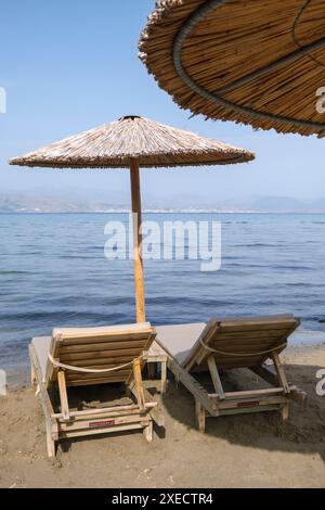 Großer Schilfschirm an einem Strand auf Korfu in Griechenland, mit ruhigem tiefblauem Meer und Liegestühlen am Sandstrand. Stockfoto