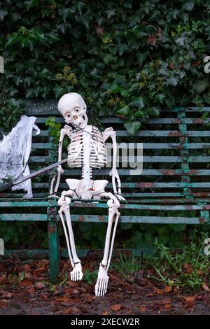 Ein Plastik-Skelett-Modell, das auf einer alten Bank sitzt, mit schrecklichen Gruselgeschenken, bereit für Haloween. Stockfoto