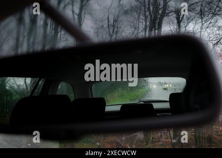 Blick durch die regenbedeckte Heckscheibe eines Autos im Fahrspiegel, während der Verkehr vorbeifährt. Stockfoto