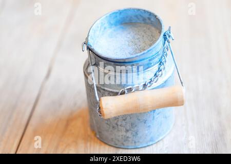 Silberner Metalleimer mit Holzgriff sitzt auf einem Holztisch. Der Eimer ist leer und hat eine Kette, die von ihm hängt. Die Szene ist einfach und unkompliziert Stockfoto