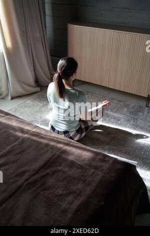 Eine Frau meditiert in einem sonnendurchfluteten Schlafzimmer, sitzt im Kreuz auf dem Boden, von hinten gefangen und schafft eine ruhige Szene. Stockfoto