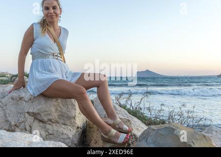 Dieses Bild fängt einen ruhigen Moment einer wunderschönen blonden Frau ein, die auf Felsen an der Küste sitzt. Sie ist elegant in einem weißen Kleid gekleidet, mit Accessoires versehen Stockfoto