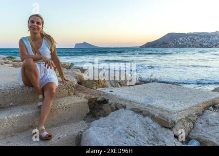Dieses Bild fängt eine schöne blonde Frau ein, die ruhig am Meer posiert. Sie ist elegant in einem weißen ärmellosen Kleid gekleidet Stockfoto