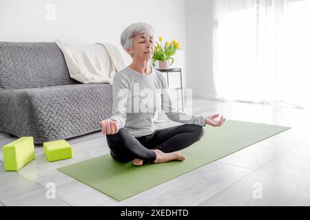 Die 70-jährige Frau praktiziert Yoga in Lotusposien zu Hause in Einem hellen Raum Stockfoto