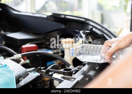 Eine Frau, die den Kühler ihres Autos auffüllt. Sie steht neben der geöffneten Motorhaube des Autos, hält eine Flasche Wasser in einer Hand und gießt sie hinein Stockfoto