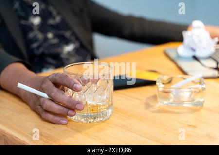 Nahaufnahme einer Hand, die ein Glas Alkohol hält. Ein zerzauster Mann sitzt in seinem Wohnzimmer, umgeben von leeren Flaschen Alkohol. Er trägt ein Knuspern Stockfoto