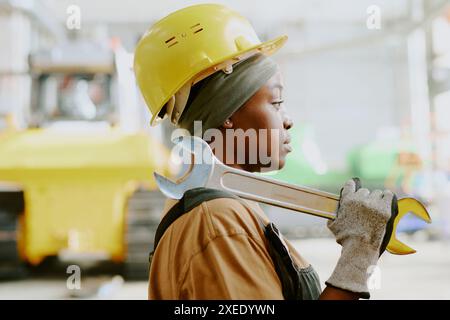 Seitliche mittlere Nahaufnahme einer jungen Afroamerikanerin, die einen Helmschlüssel trägt und am Arbeitsplatz in einer modernen Fabrik steht Stockfoto