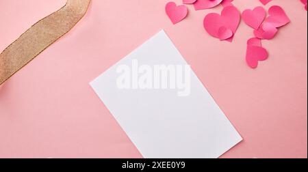 Hintergrund mit leerer Karte auf rosafarbenem Tisch mit herzförmigen Papierausschnitten und goldenem dekorativem Stoffband. Draufsicht. Stockfoto