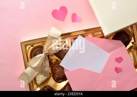 Leere Karte in einem rosafarbenen Umschlag auf Schokoladenschachtel mit goldener Schleife auf rosafarbenem Tisch mit rosafarbenen Herzausschnitten. Draufsicht. Stockfoto