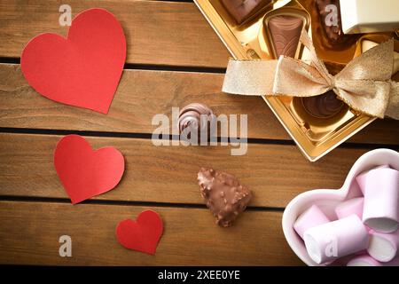 Liebhabergeschenk mit Schachtel Schokolade mit goldener Schleife und Behälter voller Süßigkeiten auf Holztisch mit Ausschnitten aus roten Herzen. Draufsicht. Stockfoto