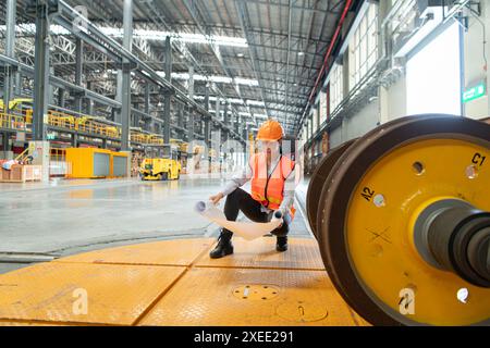 Weibliche Arbeiterin in Sicherheitsweste und Schutzhelm, die auf dem Boden in einer Skytrain-Reparaturstation sitzt und mit Blaupausen Zugräder inspiziert Stockfoto