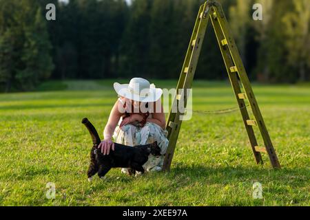 Frau auf einer Wiese mit einer Leiter, die eine Katze streichelt Stockfoto