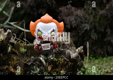 Funko Pop Actionfigur des Pennywise Evil Clown mit blutigem Skateboard aus dem Horrorfilm IT. Moody, düster, gruselig, dunkelgrüner Wald, Baumstumpf. Stockfoto