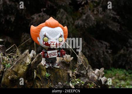 Funko Pop Actionfigur des Pennywise Evil Clown mit blutigem Skateboard aus dem Horrorfilm IT. Moody, düster, gruselig, dunkelgrüner Wald, Baumstumpf. Stockfoto