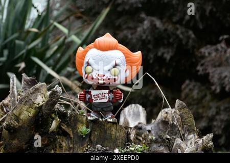 Funko Pop Actionfigur des Pennywise Evil Clown mit blutigem Skateboard aus dem Horrorfilm IT. Moody, düster, gruselig, dunkelgrüner Wald, Baumstumpf. Stockfoto