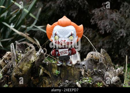 Funko Pop Actionfigur des Pennywise Evil Clown mit blutigem Skateboard aus dem Horrorfilm IT. Moody, düster, gruselig, dunkelgrüner Wald, Baumstumpf. Stockfoto