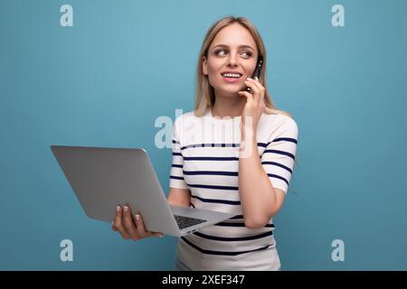 Selbstbewusste junge Geschäftsfrau mit einem Laptop in der Hand spricht am Telefon auf einem blauen isolierten Hintergrund mit Kopierraum Stockfoto